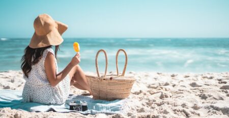 Chica en una playa, sentada en una toalla y mirando al mar. La chica lleva un vestido blanco y un gorro de paja en la cabeza.