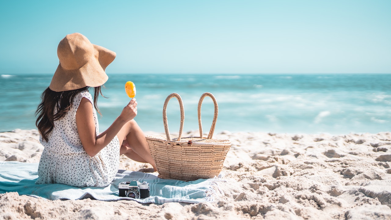 Chica en una playa, sentada en una toalla y mirando al mar. La chica lleva un vestido blanco y un gorro de paja en la cabeza.